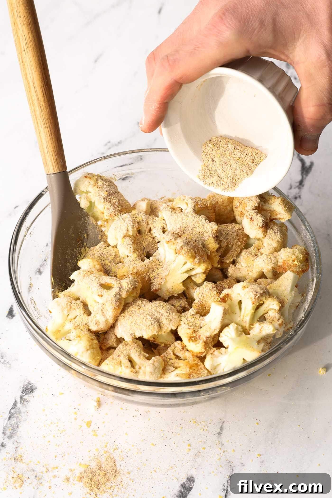 Seasoning mix being generously poured over oil-coated cauliflower florets in a bowl.
