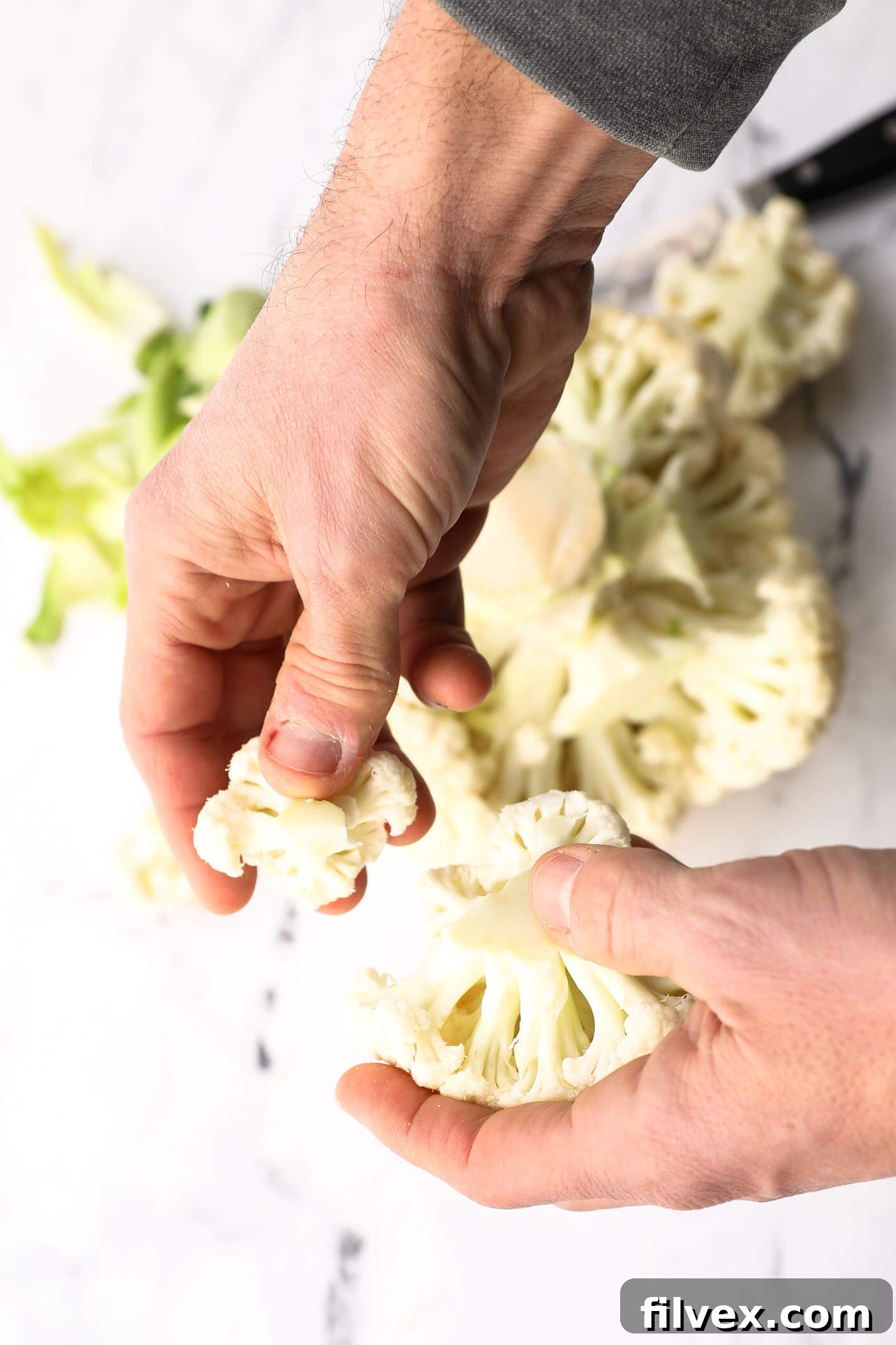 Hands breaking larger cauliflower florets into smaller, bite-sized pieces.