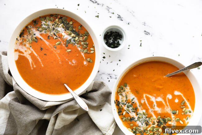 Two bowls of butternut squash and roasted red pepper soup with coconut milk drizzled on top with pumpkin seeds and chopped oregano. 
