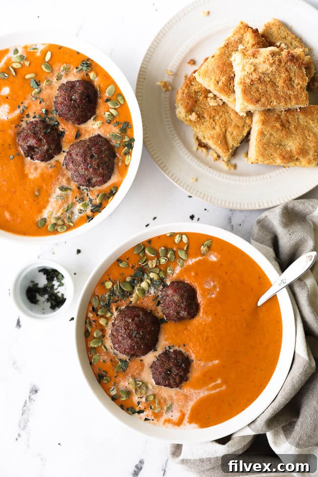 Two bowls of butternut squash and roasted red pepper soup with coconut milk drizzled on top with pumpkin seeds and chopped oregano. Served with meatballs and a side of paleo cornbread. 