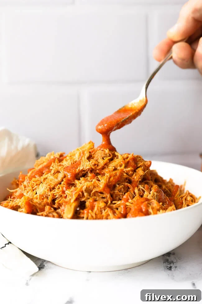 Shredded buffalo chicken in a bowl with sauce pouring in from a spoon