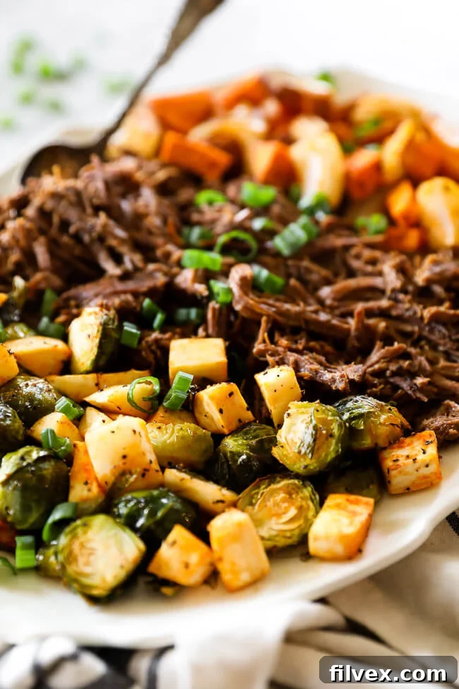 Close up angle shot of shredded slow cooker beef pot roast on platter with perfectly roasted vegetables like parsnips, sweet potatoes, and Brussels sprouts arranged on each side, highlighting the tender beef and vibrant side dishes
