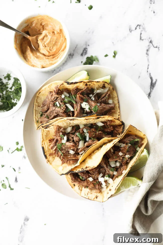 Shredded beef tacos on a plate with chopped onion and cilantro topping and sauce on the side