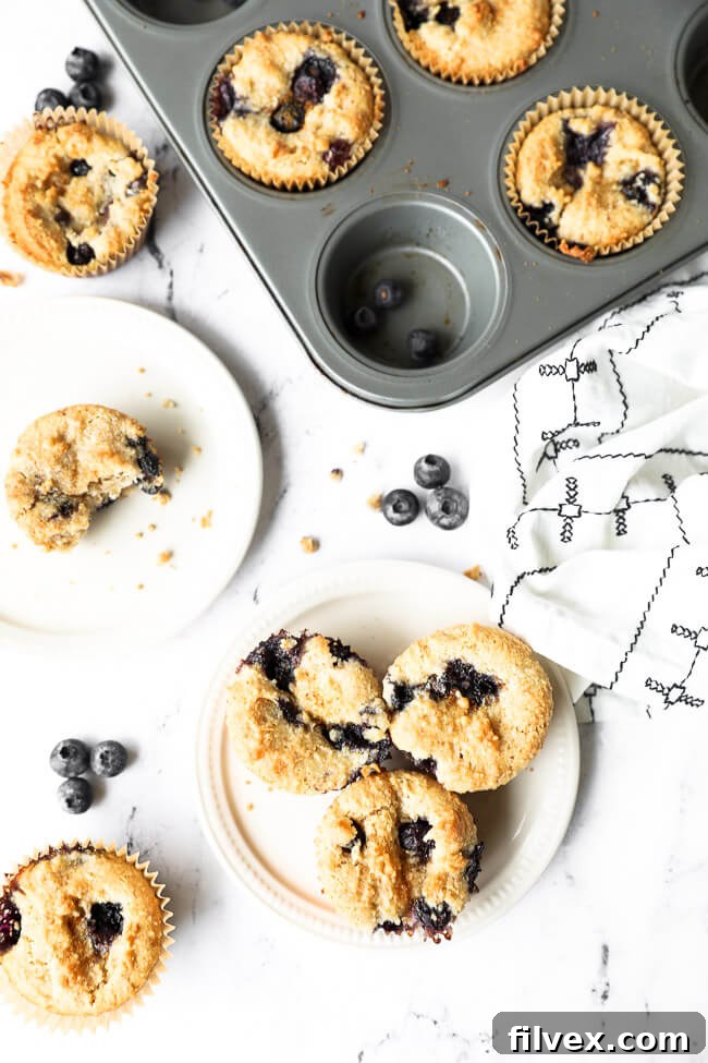 Vertical overhead image showing baked gluten-free blueberry muffins both in a baking tin and on two small plates. One plate has a single muffin with a bite taken out, revealing its soft interior.