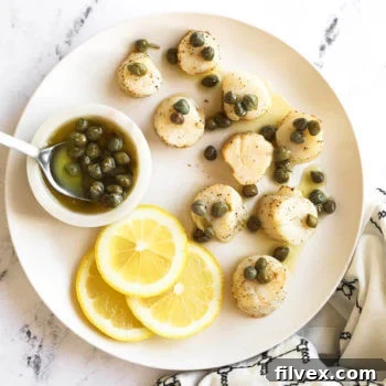 Lemon butter air fryer scallops on a white plate with butter sauce and capers on top. Lemon slices and capers in the side.