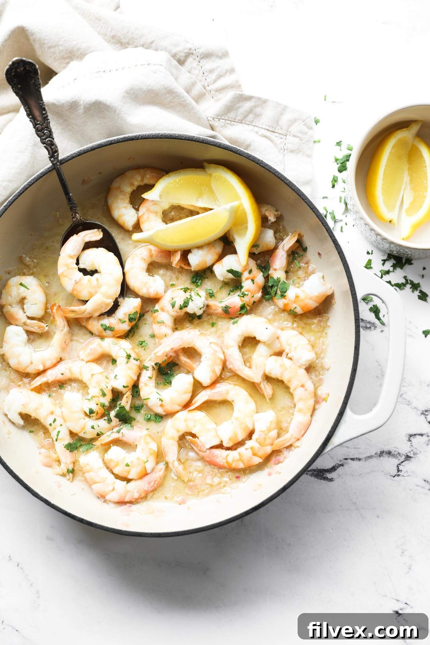 Overhead image of a skillet with garlic butter shrimp. A serving spoon dug in, and topped with chopped cilantro and lemon wedges.