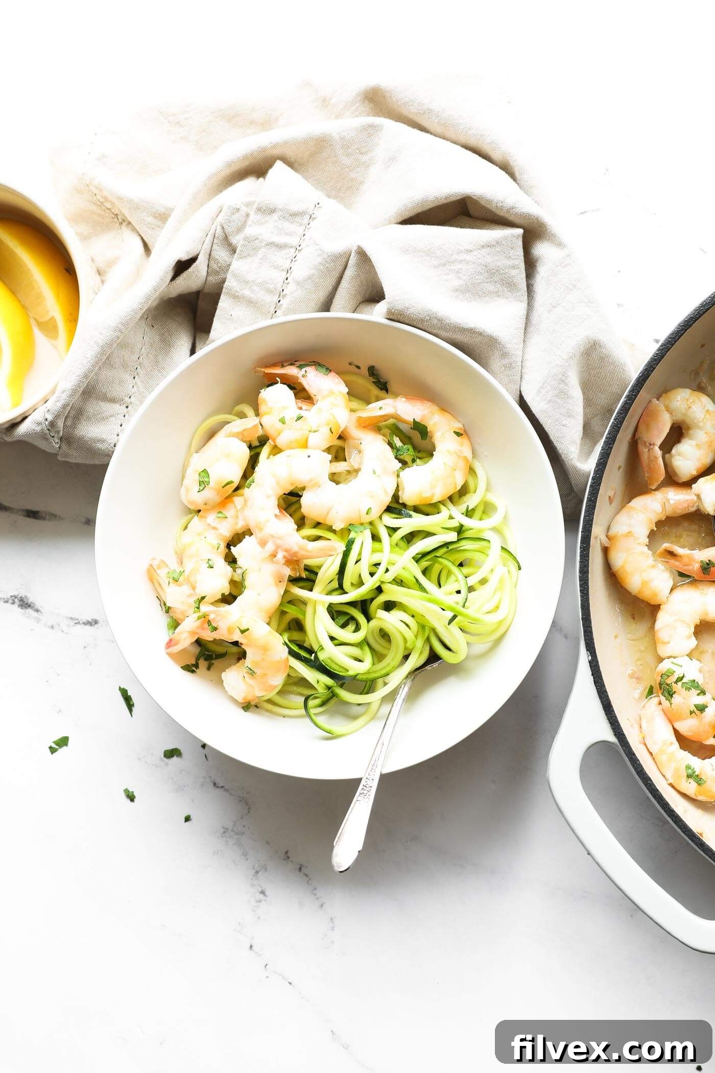 Overhead image of a garlic butter shrimp served over zoodles in a bowl with a fork. Shrimp is sprinkled with chopped cilantro on top. Extra shrimp and lemon wedges on the side.