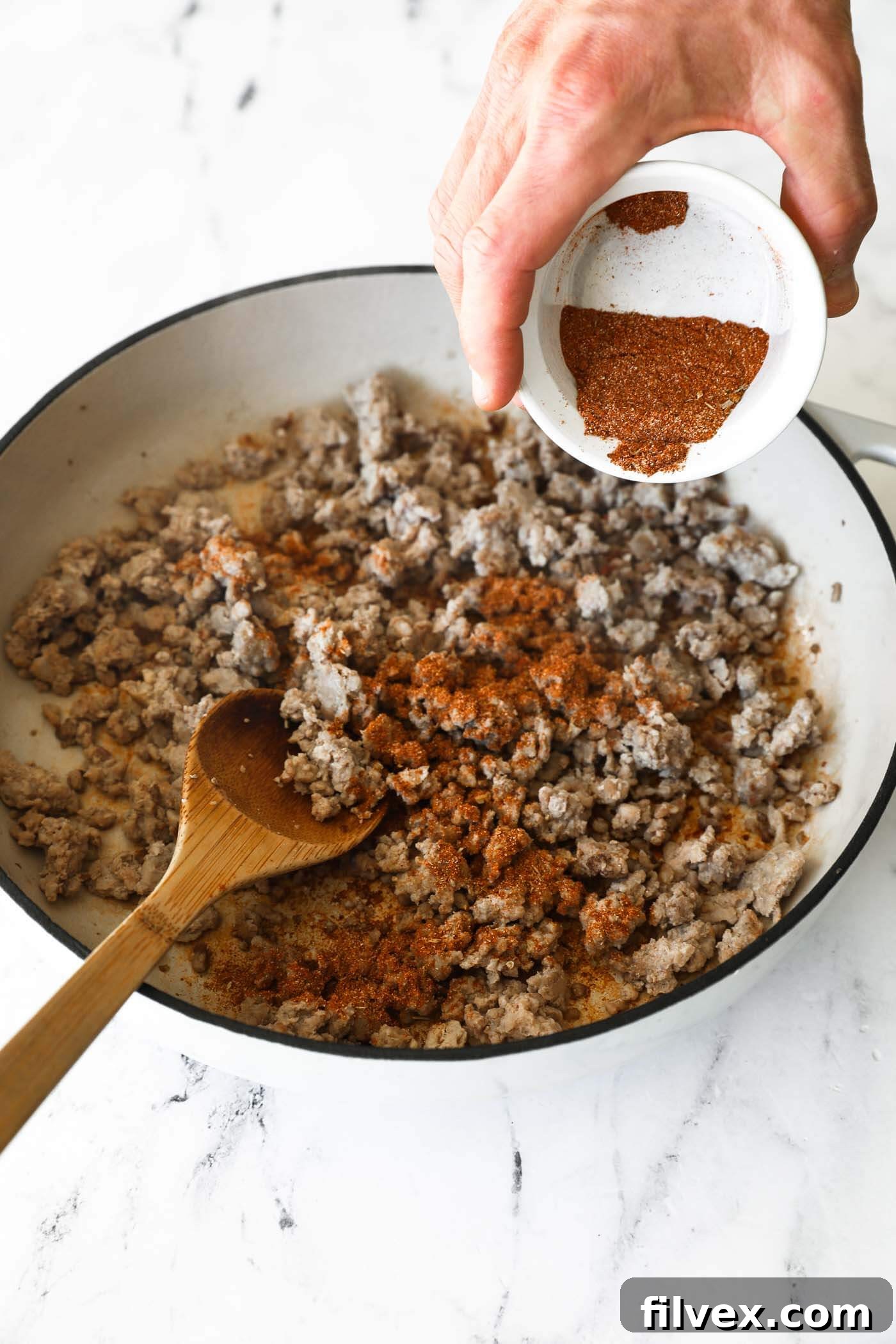 A hand stirring a generous amount of homemade taco seasoning into the cooked, crumbled ground pork in a skillet, ensuring the meat is thoroughly coated for maximum flavor.
