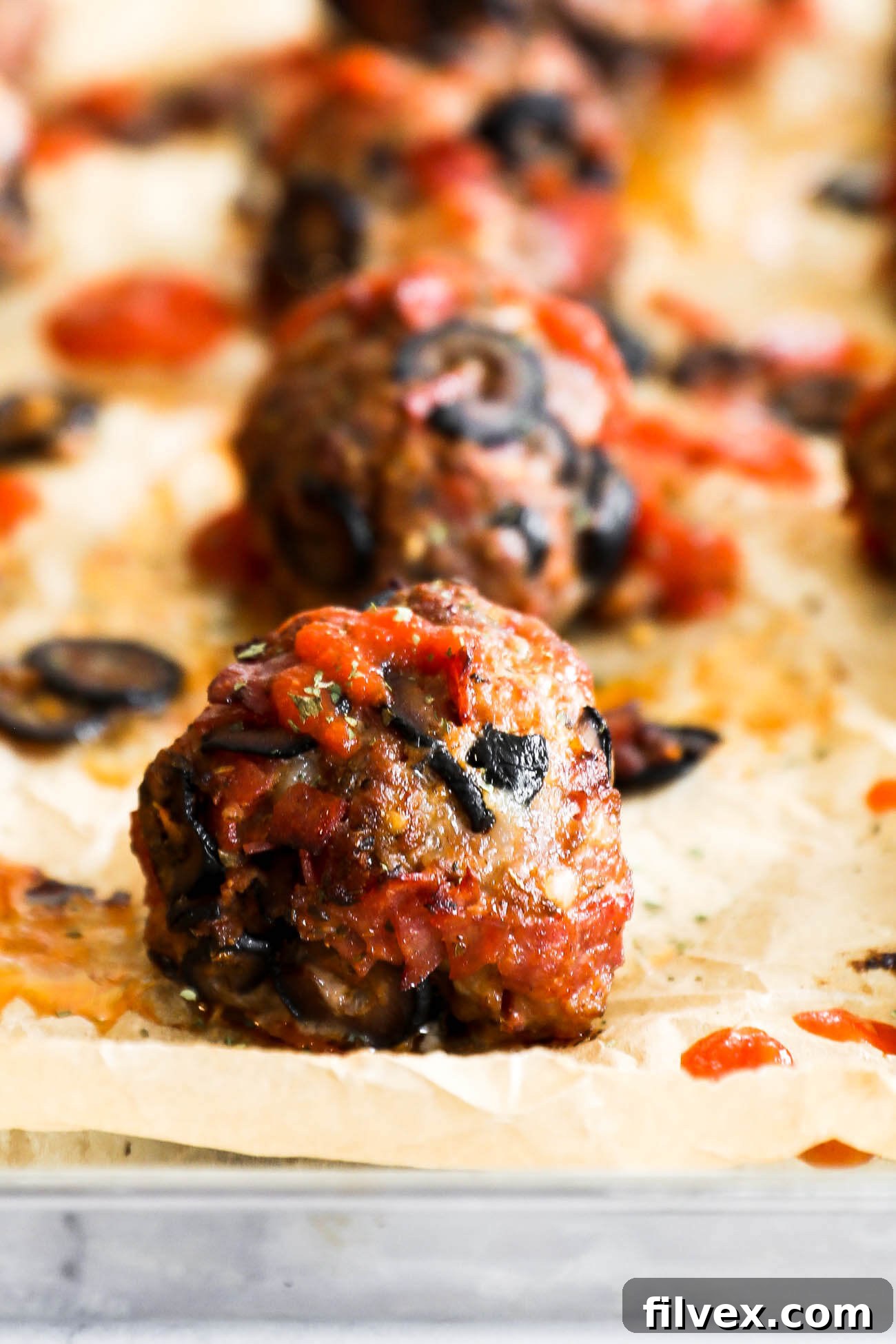 Close up of one pizza meatball on a sheet pan with more meatballs blurred out in the background. Meatball has marinara sauce drizzled on top.