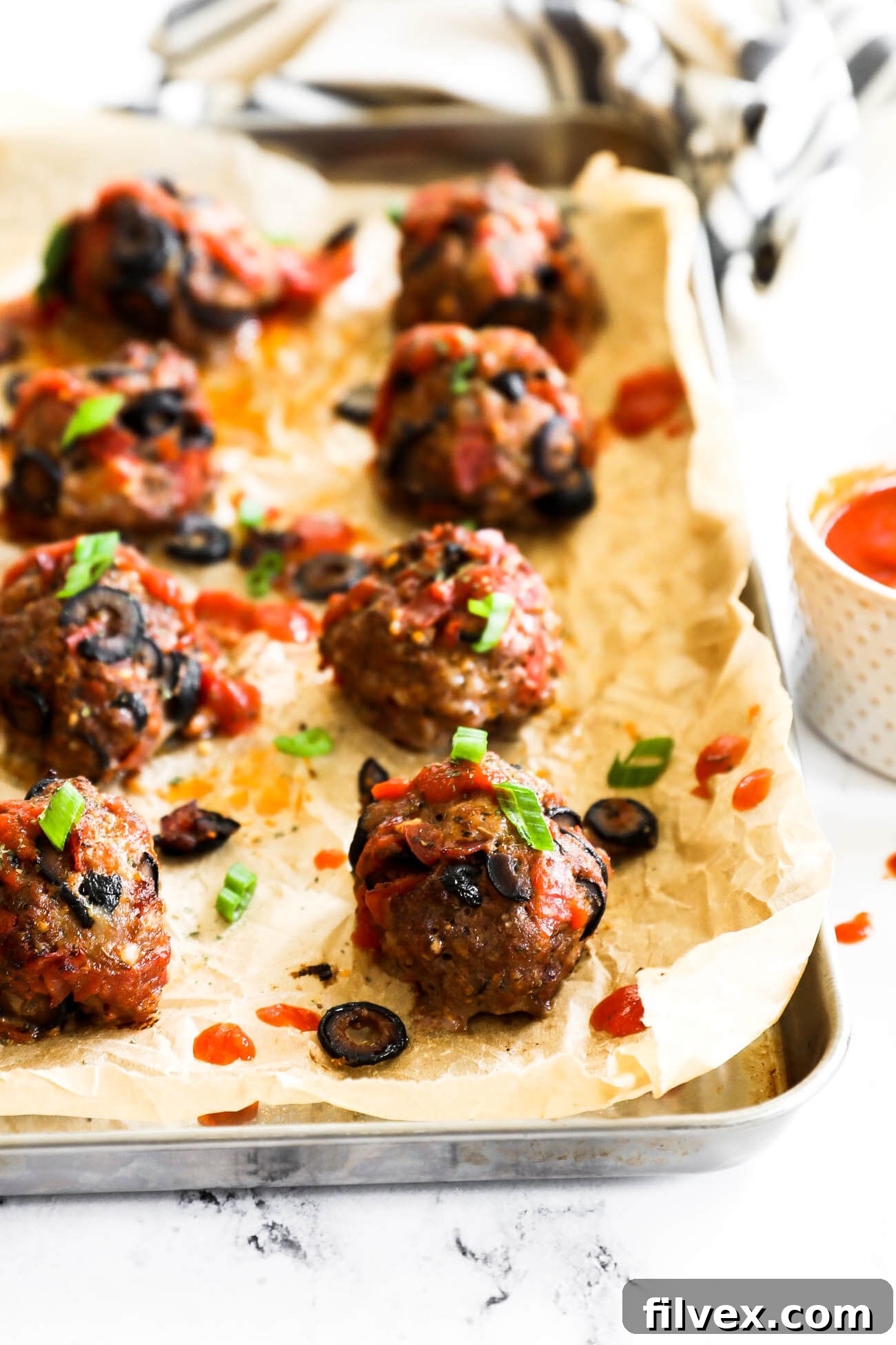 Angled image of pizza meatballs on a sheet pan with marinara sauce drizzled on top and garnished with chopped green onion.