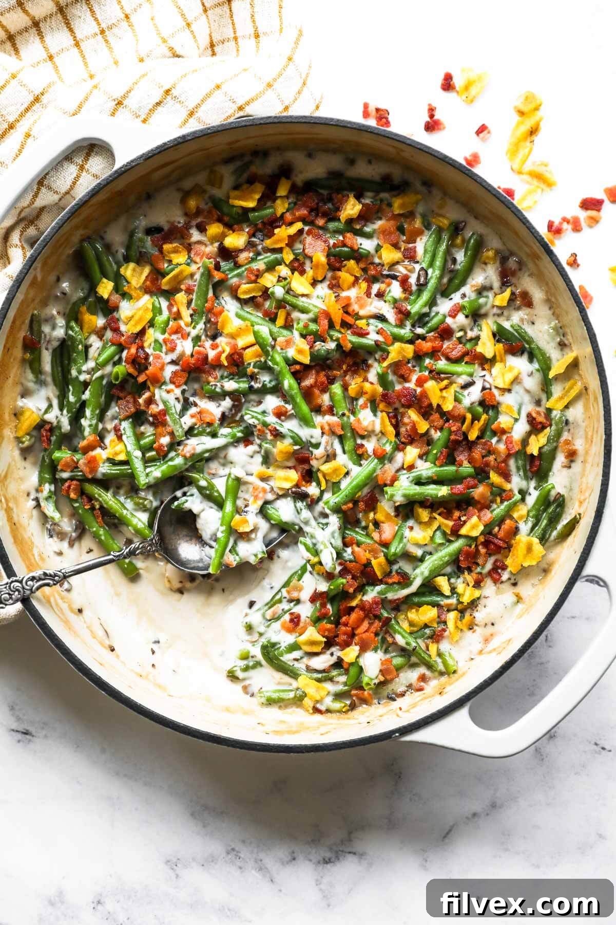 Overhead image of a skillet with green beans, a dairy free cream of mushroom sauce, crumbled bacon and plantain chips on top. Skillet has a serving spoon dug into the pan.