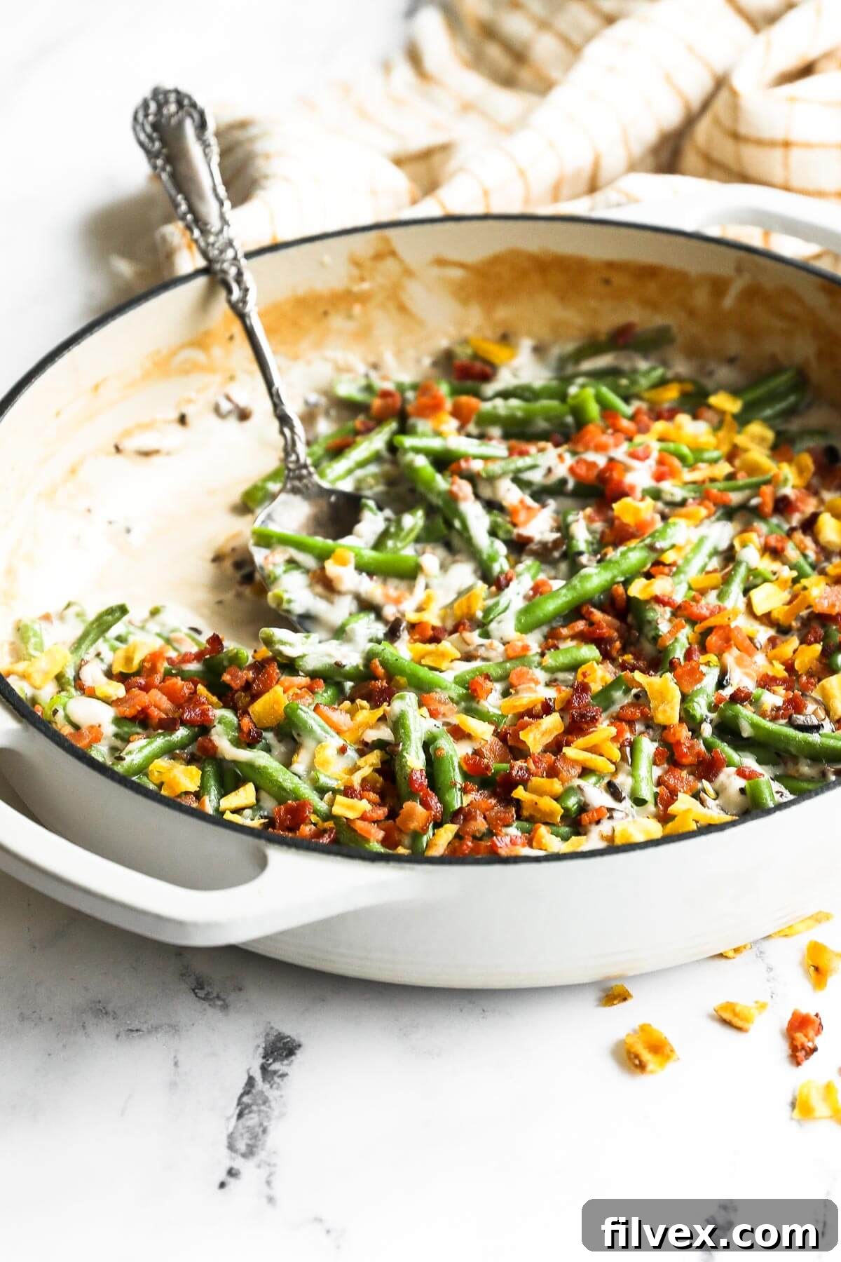 Angled image of a skillet with green beans, a dairy free cream of mushroom sauce, crumbled bacon and plantain chips on top. Serving spoon dug into the casserole.