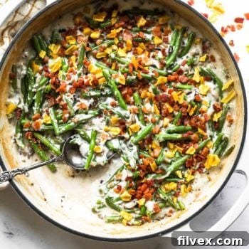 Overhead image of a skillet with green beans, a dairy free cream of mushroom sauce, crumbled bacon and plantain chips on top. Skillet has a serving spoon dug into the pan.