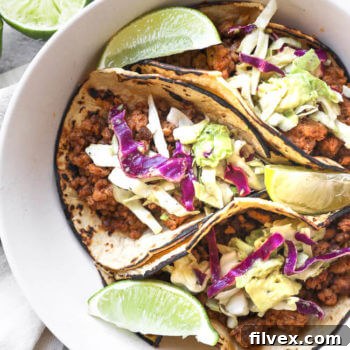 Three perfectly assembled ground pork tacos with a fresh cabbage and avocado slaw, served in a bowl with vibrant lime wedges for garnish. This appetizing image highlights the rich textures and flavors of a simple yet satisfying meal.