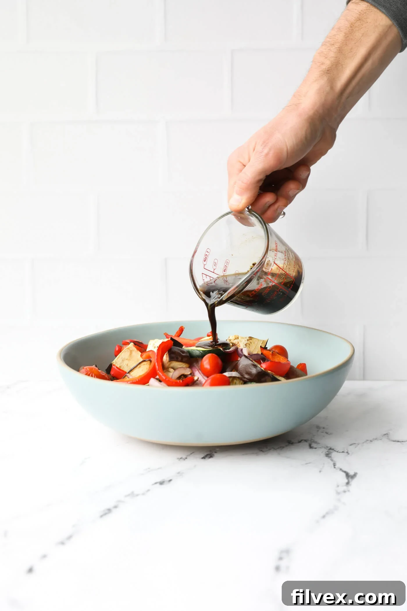 A hand pouring the prepared balsamic marinade over the hot grilled vegetables in a bowl.