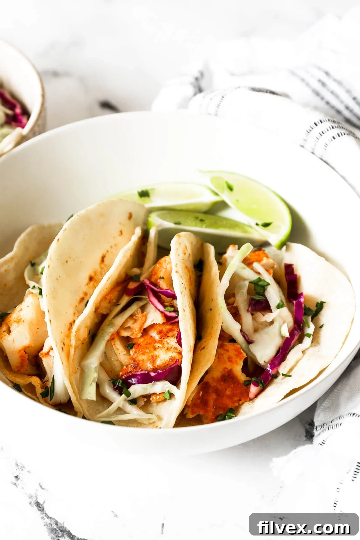 Close up angled image of a bowl with three fish tacos made in the air fryer. Lime wedges and chopped cilantro garnish.