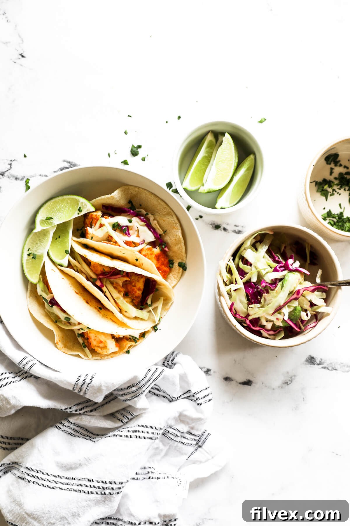 Overhead image of a bowl with three fish tacos and smaller bowls with lime wedges, a citrusy coleslaw and chopped cilantro all on the side.