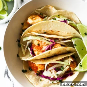 Flatlay image of three fish tacos situated in a bowl with a few lime wedges and chopped cilantro on top.