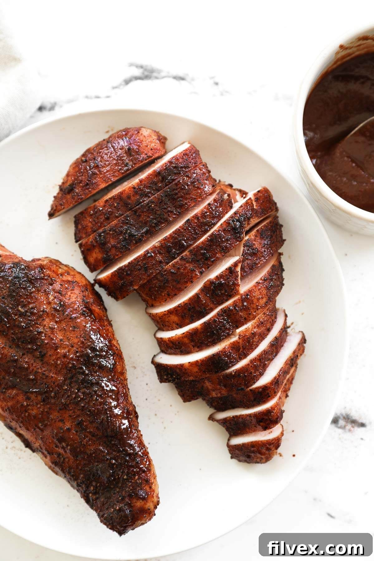 Two pieces of smoked chicken breast on a plate, one sliced, with a small bowl of BBQ sauce on the side.