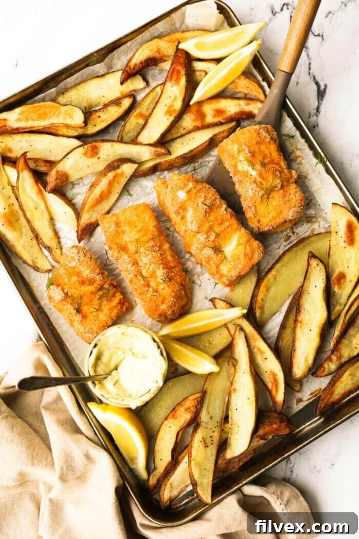 Overhead image of a sheet pan with golden-brown baked fish and crispy potato wedges, garnished with fresh lemon slices and a jar of homemade tartar sauce, ready to be served.