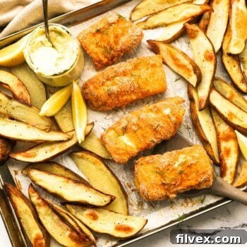 Overhead image of a sheet pan with golden baked fish and chips. The crispy battered fish pieces are nestled amongst baked potato wedges. A jar of homemade tartar sauce and lemon wedges complete the garnish.
