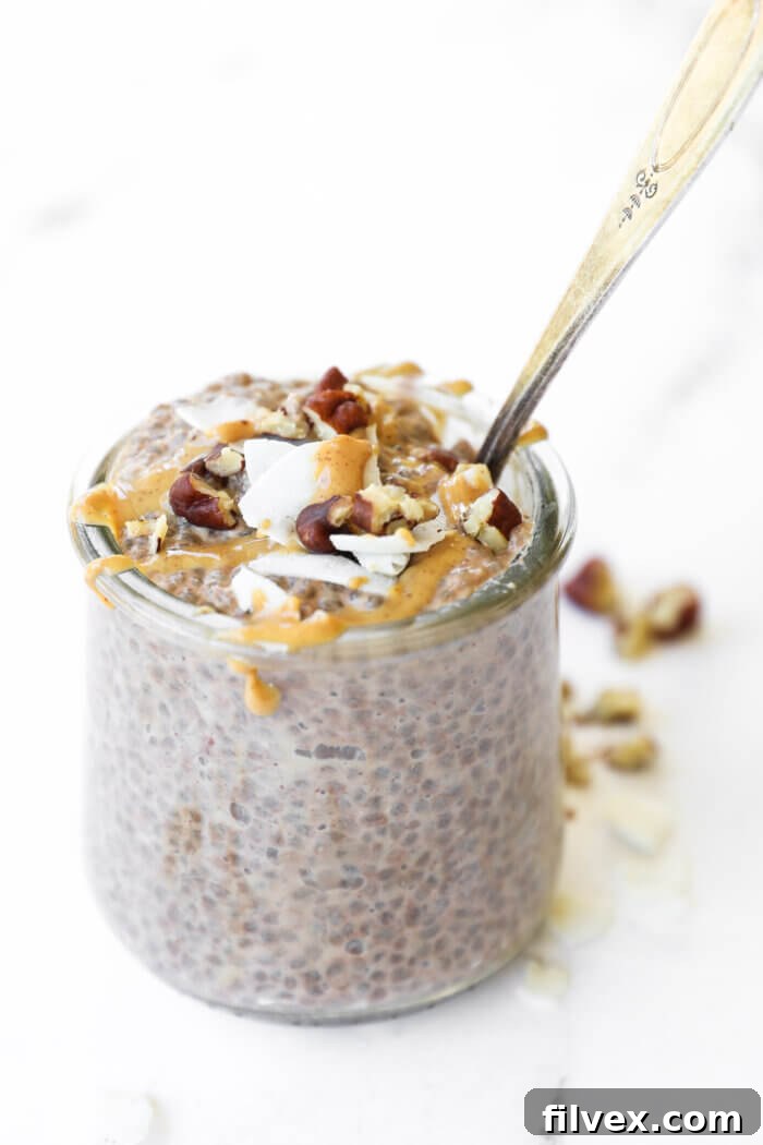 A small glass jar filled with luscious peanut butter chocolate keto chia pudding, topped generously with fresh raspberries, chopped pecans, and a drizzle of peanut butter. A spoon is poised, ready for a delicious bite.