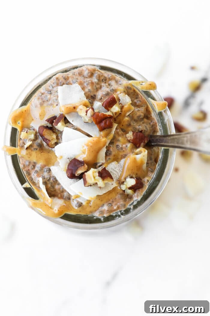 Close-up overhead view of keto chia pudding in a clear mason jar, generously topped with a dollop of peanut butter, a sprinkle of chopped pecans, and delicate coconut flakes.