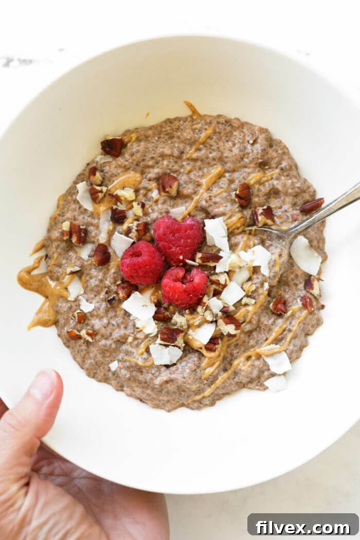 Close-up shot of a hand holding a bowl of peanut butter chocolate keto chia pudding, adorned with a variety of toppings including shredded coconut, crunchy chopped pecans, a drizzle of creamy peanut butter, and a scattering of fresh raspberries. A spoon is ready to scoop.