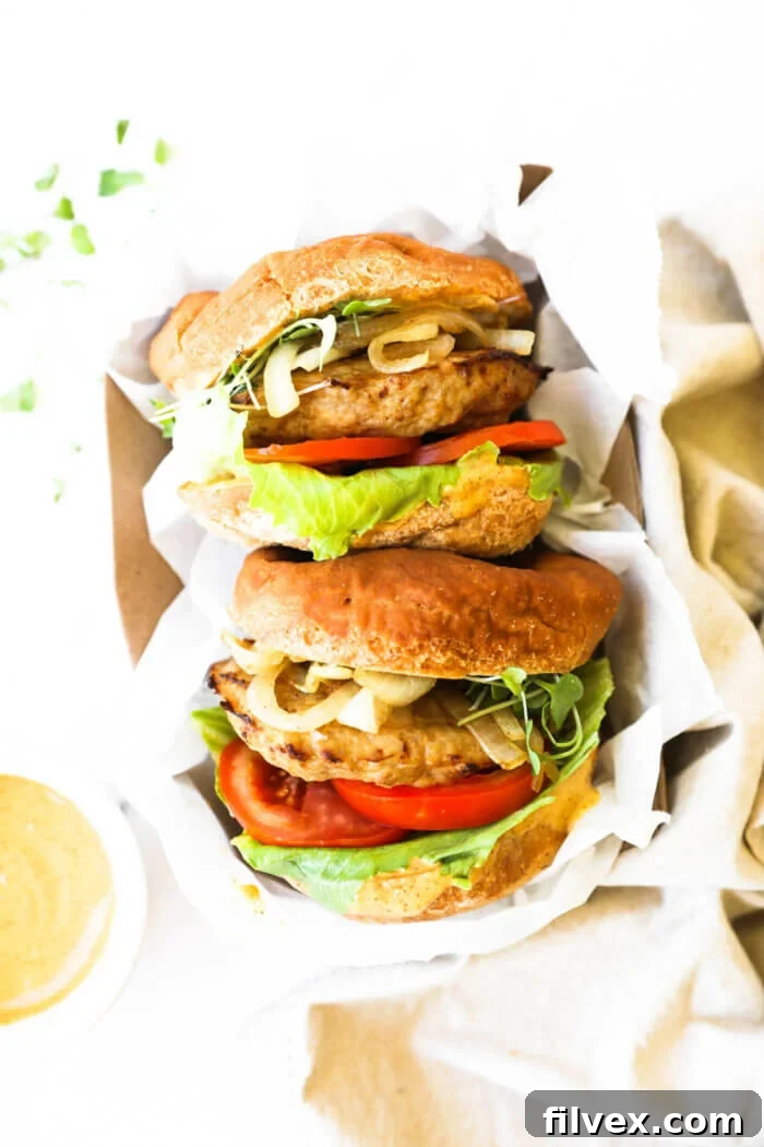 Two fully loaded turkey burgers in a convenient to-go container lined with parchment paper, accompanied by a side of chipotle aioli sauce for dipping.