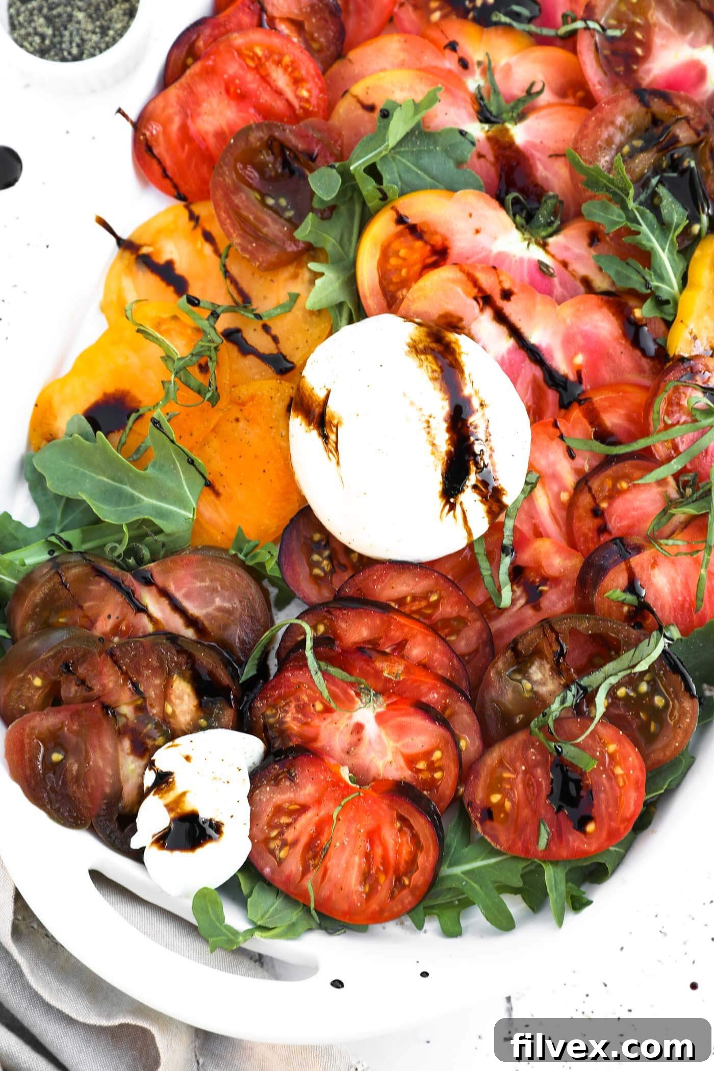 Close up overhead image of sliced heirloom tomatoes on a bed or arugula lettuce. Topped with burrata cheese, basil and drizzled with balsamic vinegar.