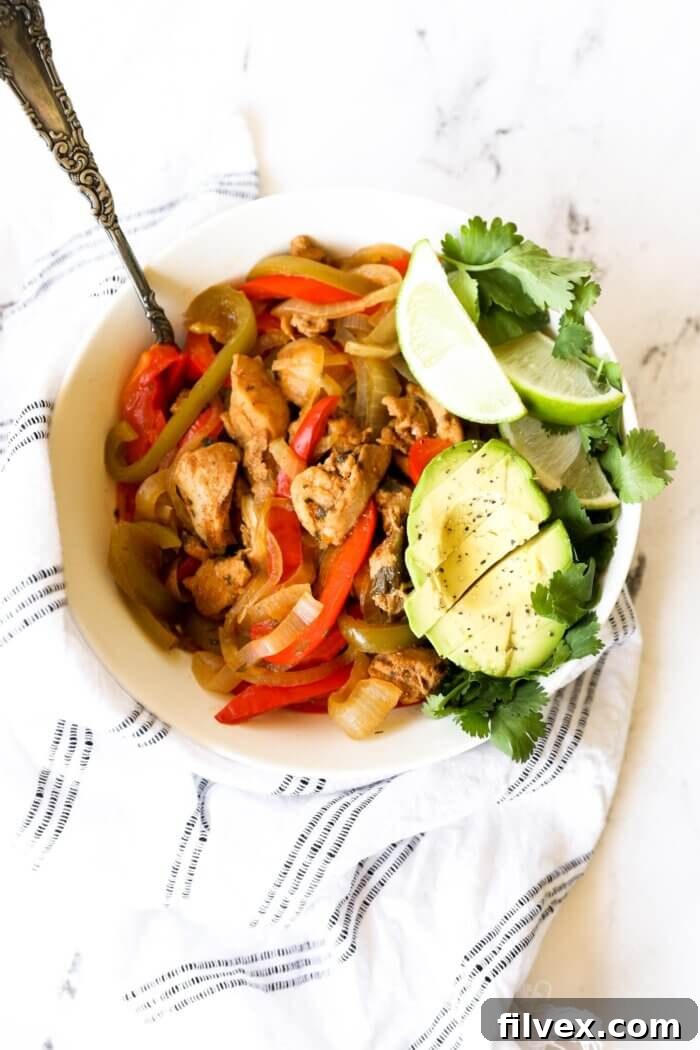 Overhead image of a bowl full of chicken fajitas. Garnished with half an avocado sliced, lime wedges and fresh cilantro. A serving spoon is dug into the bowl, inviting you to enjoy.