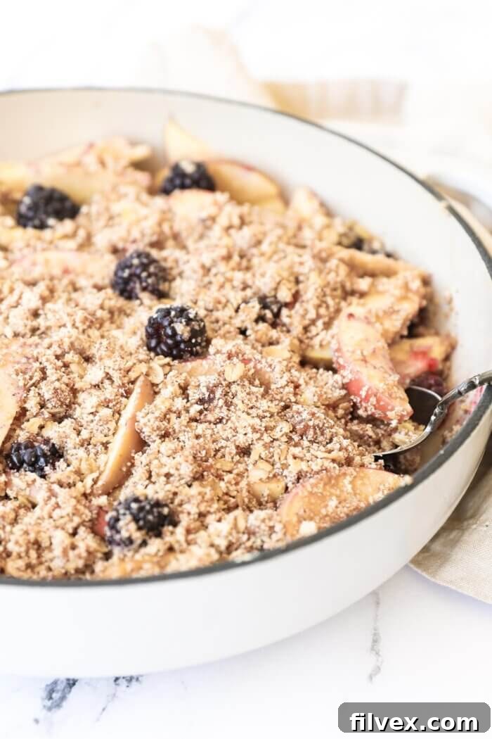 Angled image of blackberry and apple crumble in a skillet to show the crumbly texture of the topping, with a spoon revealing the juicy fruit filling beneath.