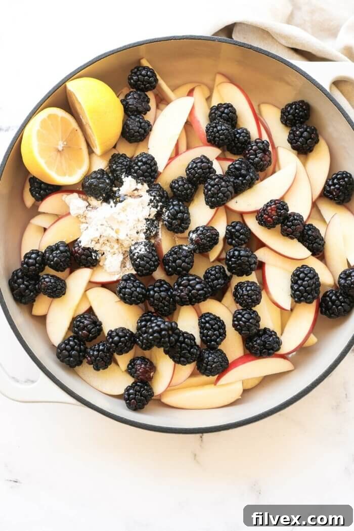 Overhead image of skillet full of apple and blackberry crumble filling ingredients, including apple slices, blackberries, tapioca starch, maple syrup and 2 lemon halves, all ready to be mixed.