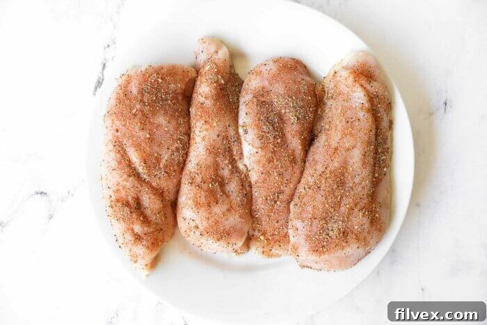 Four raw chicken breasts generously coated in the seasoning mix on a plate, ready for the next step.