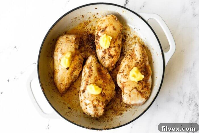Beautifully browned chicken breasts in a skillet, each topped with a pat of butter, after the initial sear.