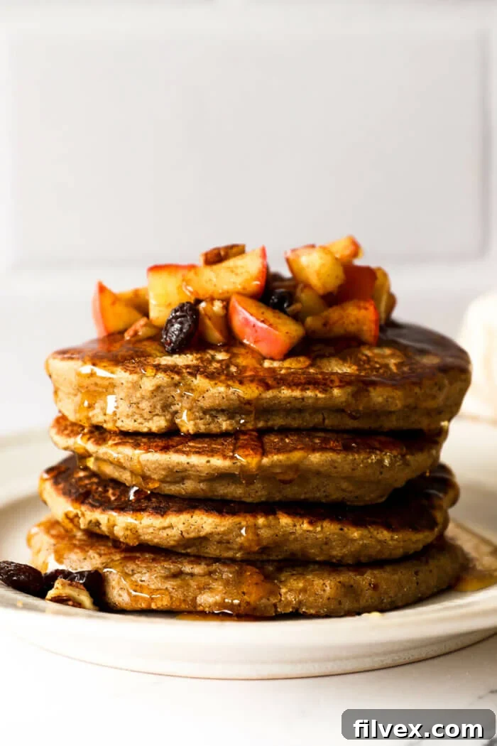 A stack of golden-brown oat flour pancakes, generously topped with sautéed apples, raisins, and pecans, all drizzled with rich maple syrup.