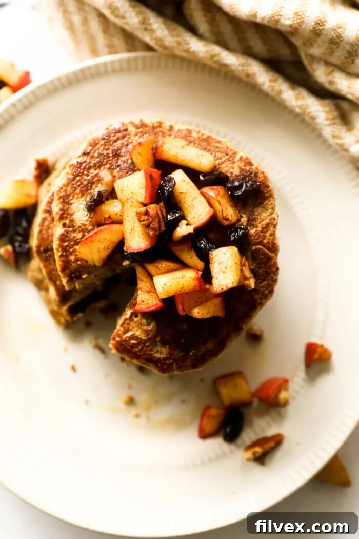 An overhead view of a luscious stack of oat flour pancakes, adorned with a vibrant apple, raisin, and pecan topping, glistening with maple syrup.