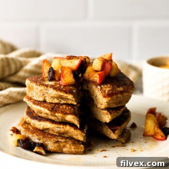 A stack of oat flour pancakes on a plate with one slice removed. They are topped with sauteed apples, raisins and pecans, and drizzled with maple syrup.
