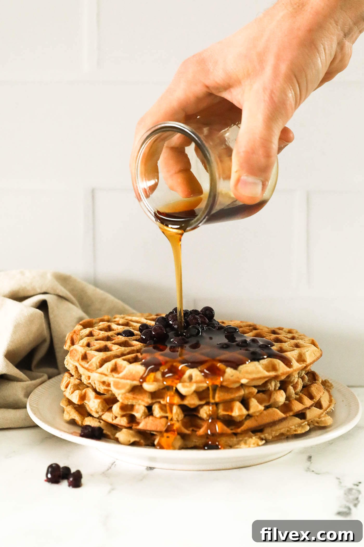 Image of a hand pouring maple syrup on a stack of waffles with wild blueberries on top. Golden, crispy gluten-free oat flour waffles.