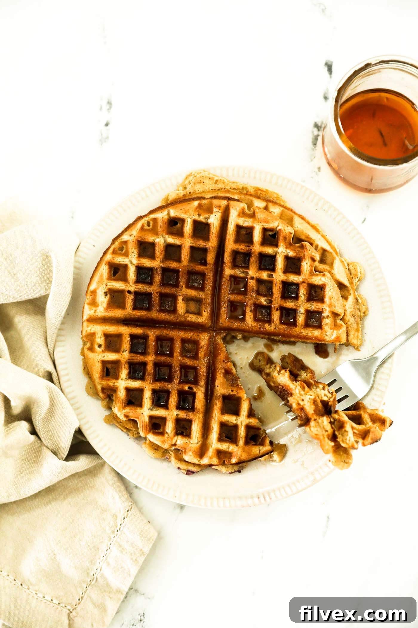A plate with oat flour waffles. A triangle bite has been cut out and is on the fork next to the rest o the waffle. Maple syrup has been poured on top and more is on the side, highlighting the crispy exterior and fluffy interior.