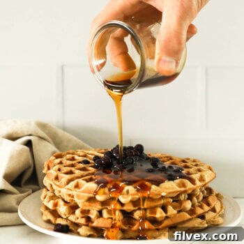 Image of a hand pouring maple syrup on a stack of waffles with wild blueberries on top.