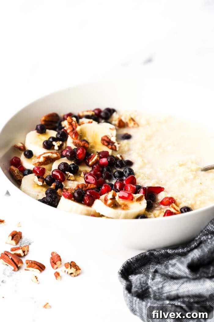 Angled image of a bowl of creamy millet porridge with coconut milk, bananas, berries, pomegranate seeds and chopped pecans, highlighting the texture and fresh toppings.