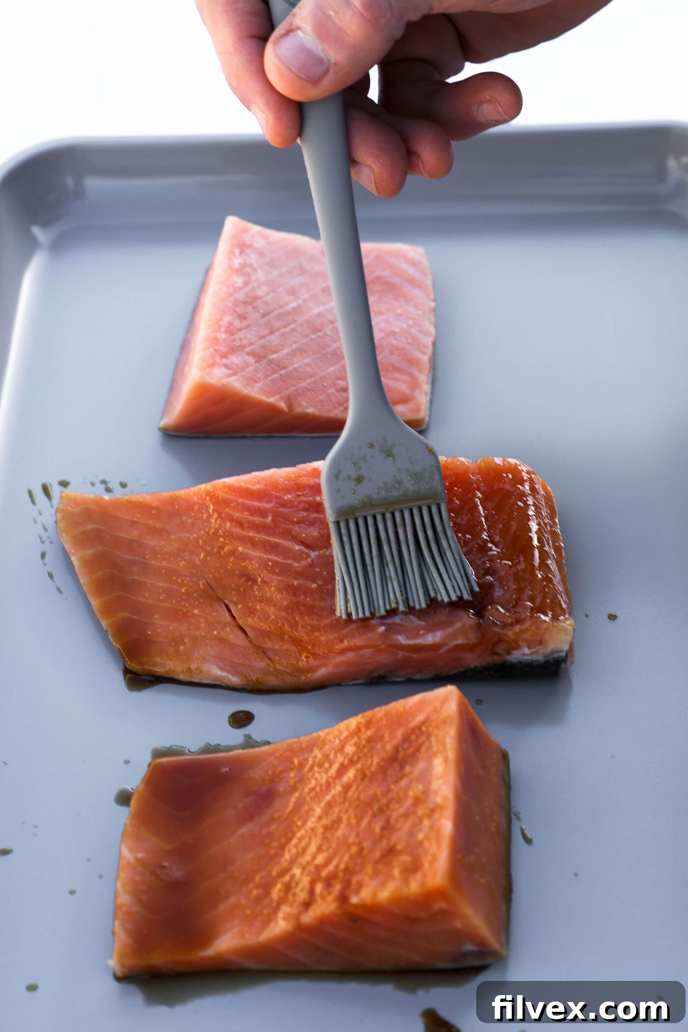 A pastry brush applying the glaze generously over a salmon portion on a sheet pan.