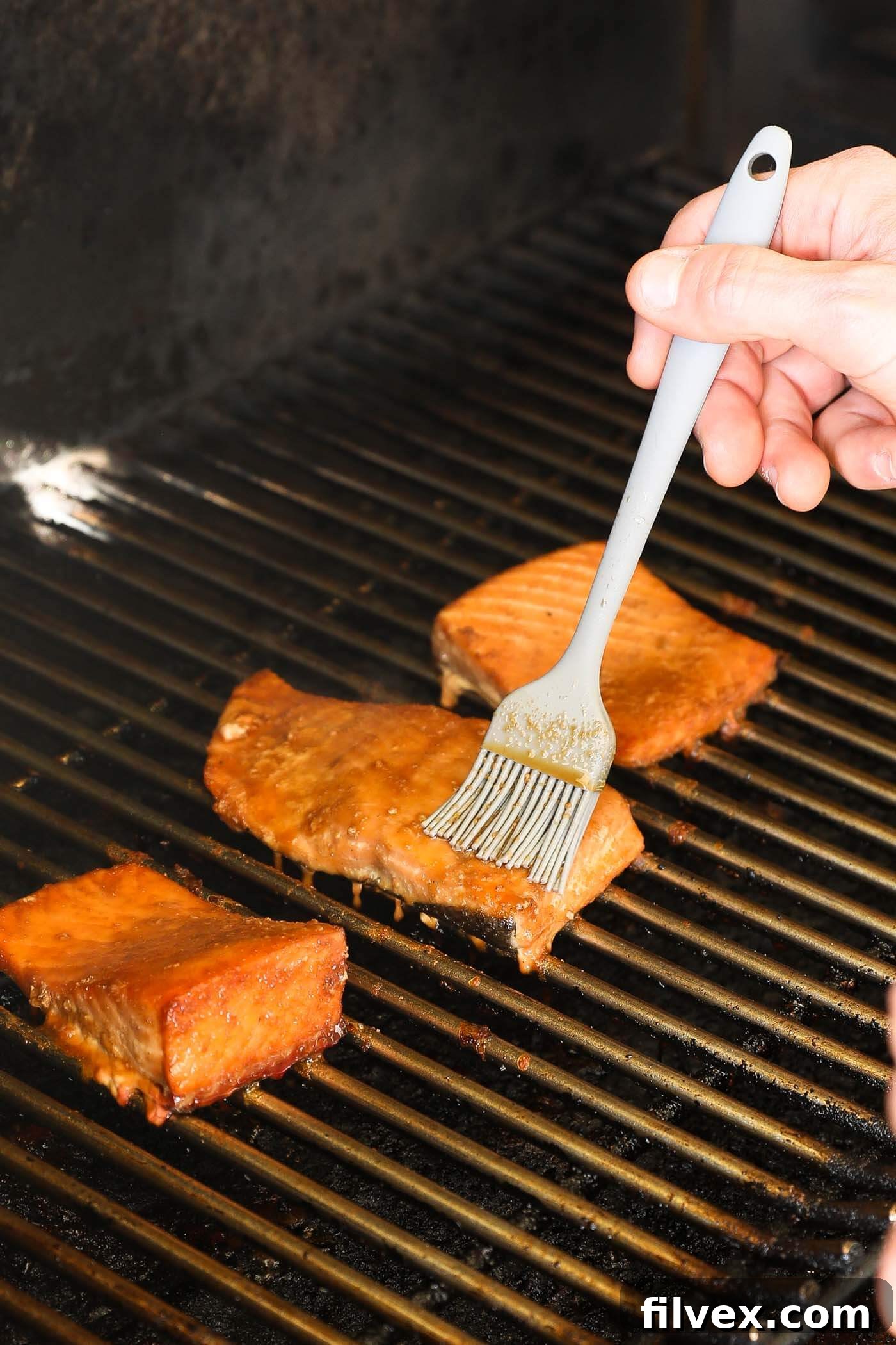 A hand brushing more glaze onto a smoking salmon fillet on a grill.