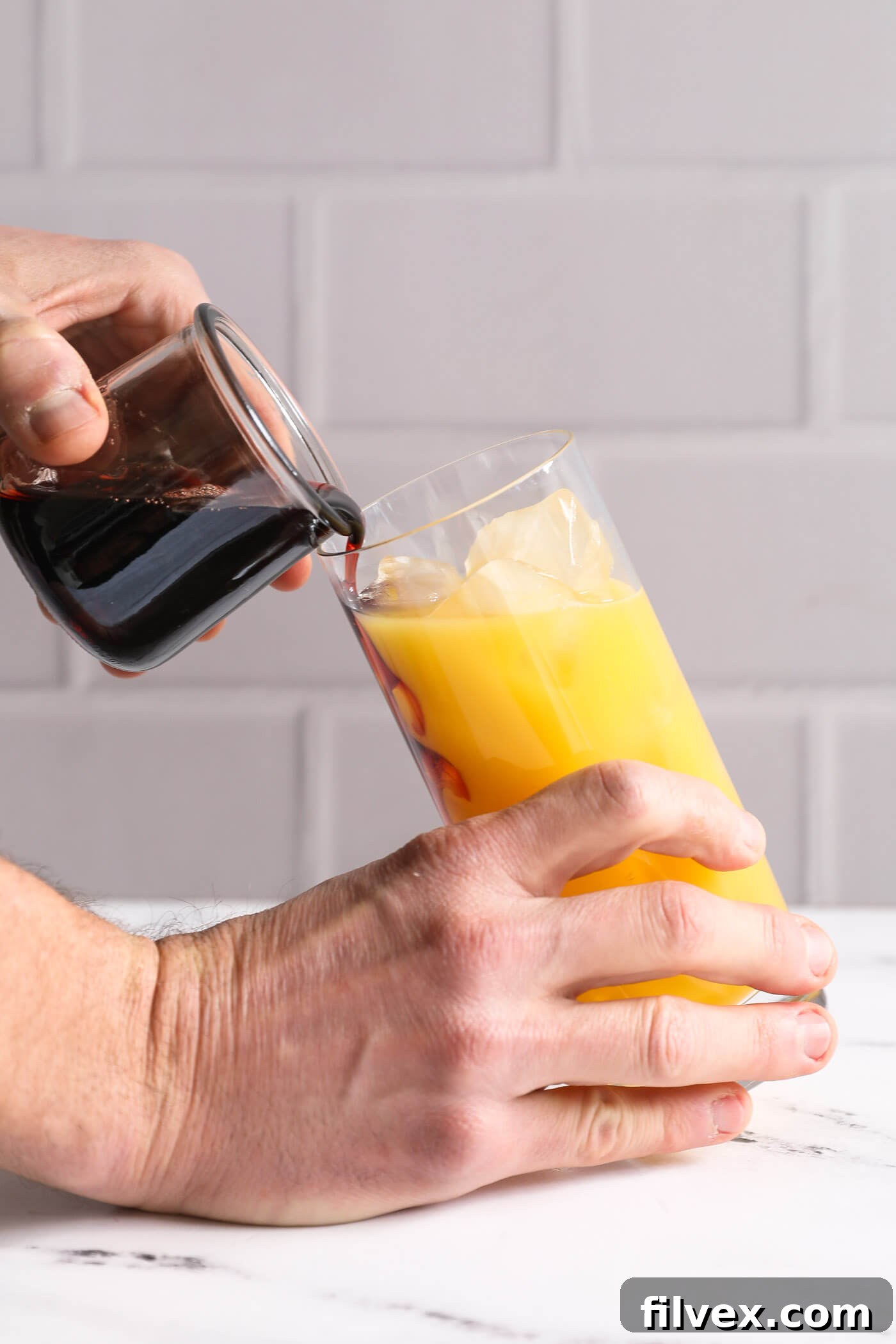 Grenadine syrup being carefully streamed down the inside wall of a tilted glass to create a perfect, distinct red layer at the bottom of the orange juice.