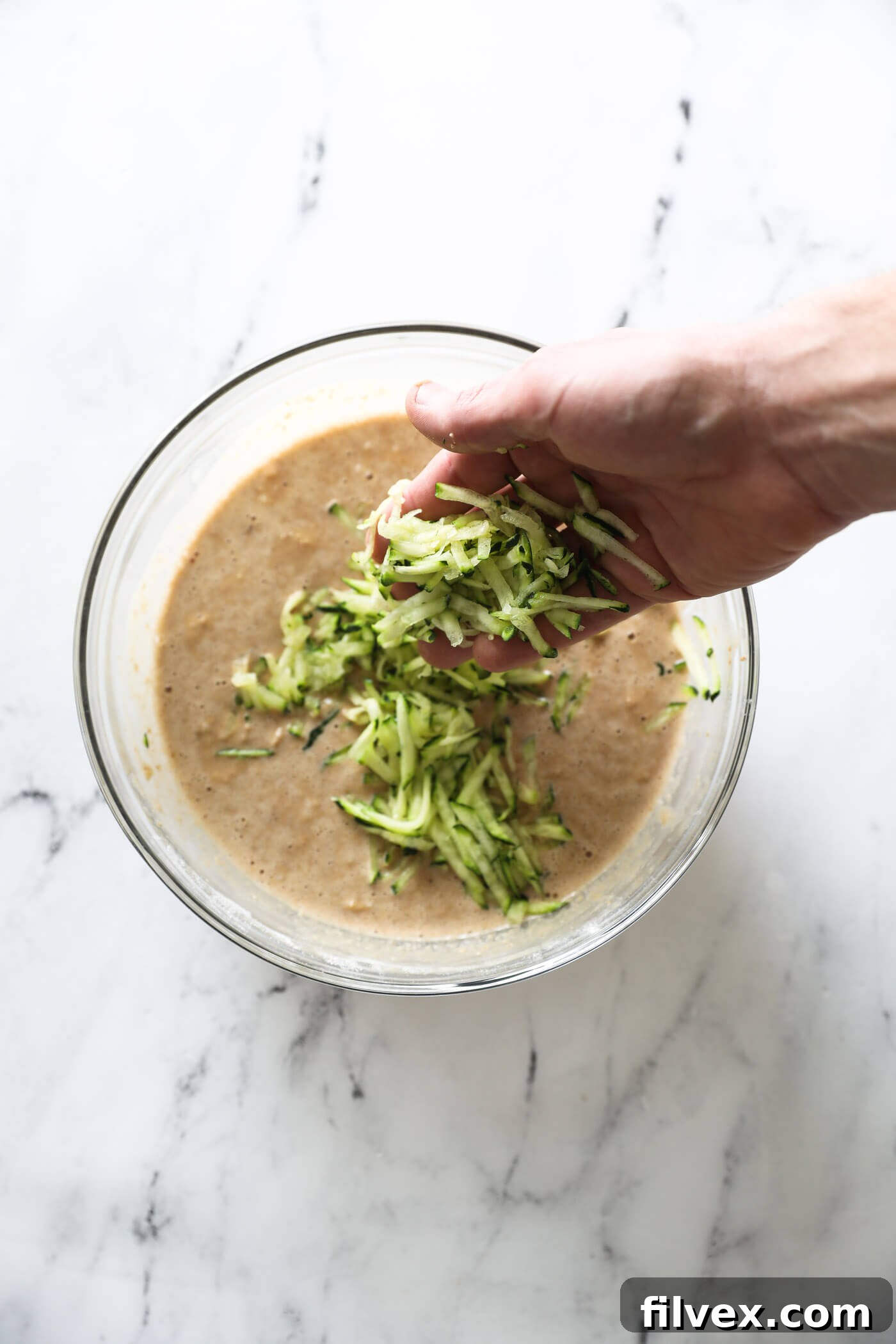 A hand adding the shredded zucchini to the batter in a mixing bowl.
