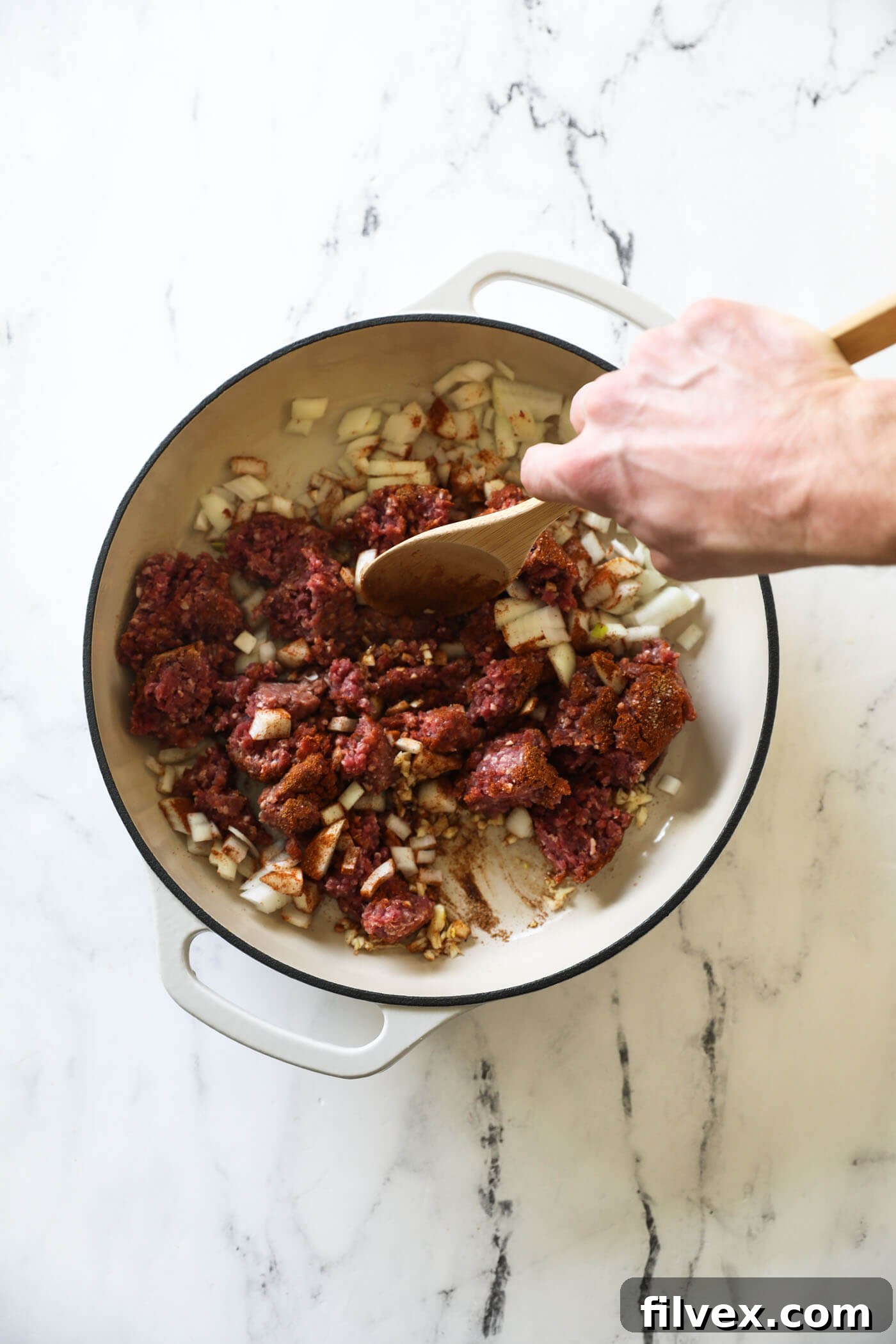 A close-up view of ground beef in a pan being broken into smaller, uniform chunks with a wooden spoon, ensuring it browns evenly with the onions and garlic.