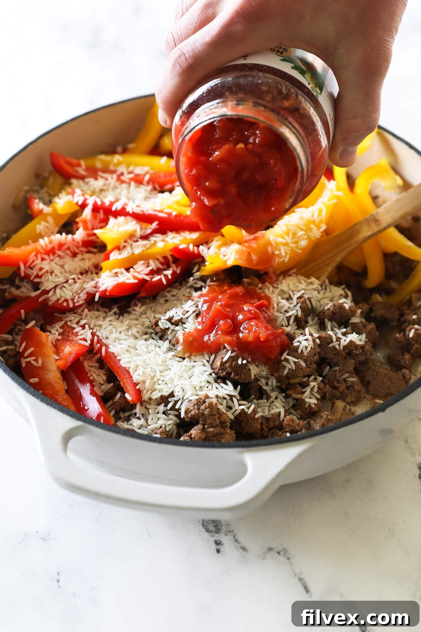 The browned ground beef mixture in the pan, with sliced bell peppers, a can of diced tomatoes, uncooked rice, and water freshly added, ready to be stirred.
