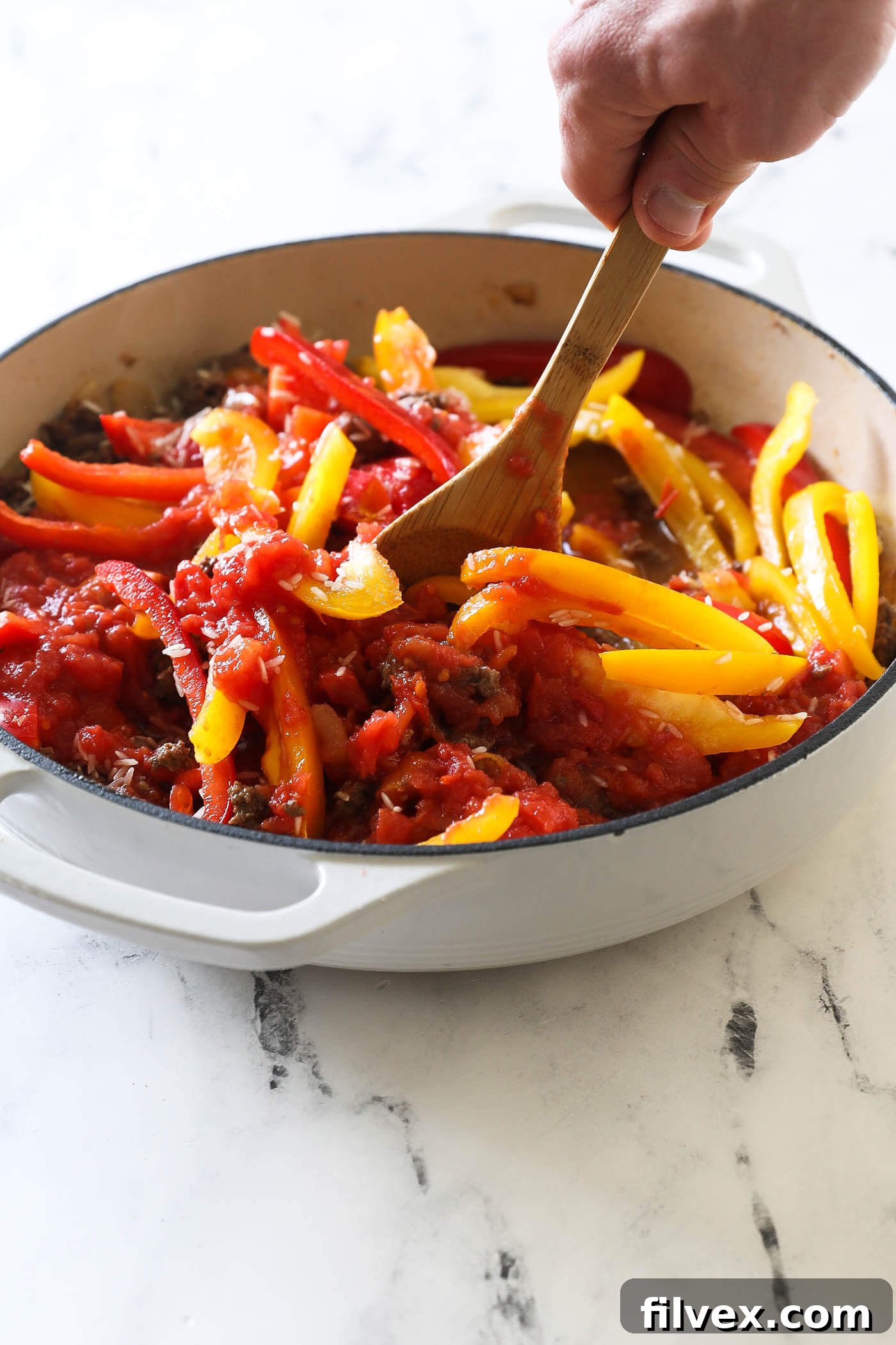 A wooden spatula vigorously stirring the mixture of ground beef, rice, bell peppers, diced tomatoes, and water in a large pan, ensuring all components are well-integrated.