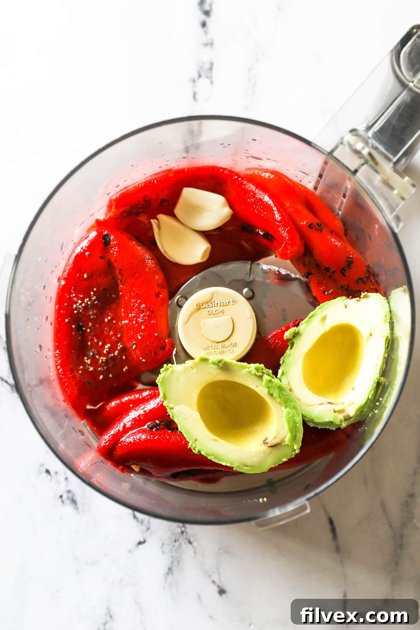 All three main ingredients - drained roasted red peppers, a ripe avocado, and garlic cloves - placed in a food processor, ready for blending.